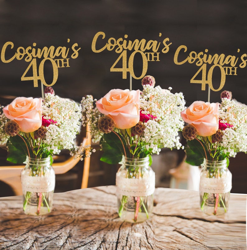 May include: Three gold glitter table decorations with the text "Cosima's 40th" on each. The decorations are placed in front of three mason jars filled with flowers.