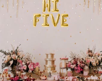 May include: A pink and gold dessert table with the words "Hi Five" in gold balloons. The table is decorated with flowers, candles, and other festive decorations.