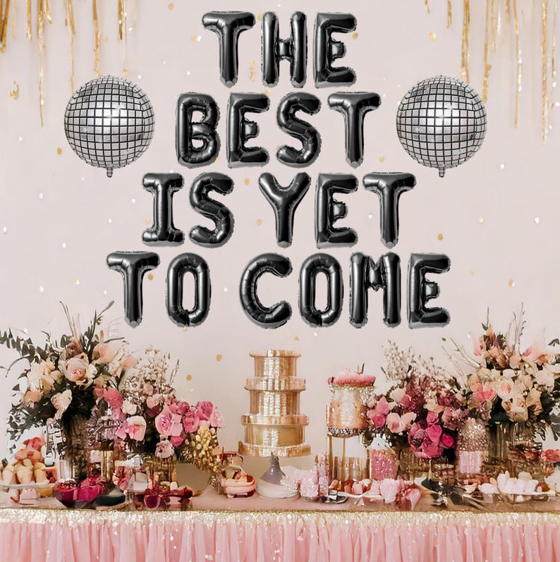 May include: Black balloon letters spelling "The Best Is Yet To Come" with silver disco ball decorations on a white wall. A table with a pink tablecloth is decorated with flowers and cake stands.