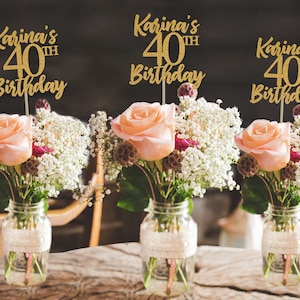 Puede incluir: Tres centros de mesa de purpurina dorada con el texto "Karina's 40th Birthday" en cada uno. Los centros de mesa est&aacute;n colocados en tarros de cristal con flores.