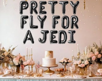 May include: Black balloon letters spelling "Pretty Fly For A Jedi" on a pink wall above a table with a white cake and floral arrangements.