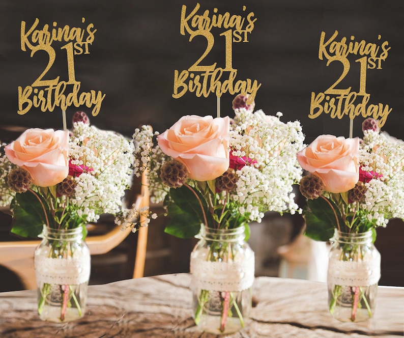 Puede incluir: Tres centros de mesa de purpurina dorada con el texto "Karina's 21st Birthday" en cada uno. Los centros de mesa se colocan en tarros de vidrio transparentes con encaje alrededor de la parte superior y se llenan con flores blancas y rosas.
