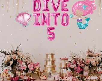 May include: Pink and blue mermaid themed party decorations with the words "Dive Into 5" in pink balloons. The table is decorated with pink and white flowers and a pink tablecloth.