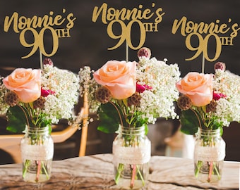 May include: Three gold glitter table centerpieces with the text "Nonnie's 90th" on each. The centerpieces are placed in mason jars with lace around the top and filled with white and pink flowers.