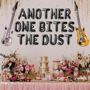 May include: Black balloon letters spelling "Another One Bites The Dust" with two gold electric guitar shaped balloons on either side. The balloons are hanging on a white wall above a table with pink and white flowers and a tiered cake stand.
