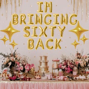 May include: Gold foil balloons spelling "I'm Bringing Sixty Back" with gold star balloons on a white wall. A table with a pink tablecloth is decorated with flowers, desserts, and a tiered cake stand.