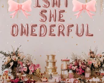May include: A party scene with pink bow-shaped balloons and rose gold foil letter balloons spelling "ISNT SHE ONEDERFUL." A table is laden with desserts and floral arrangements, creating a festive atmosphere.