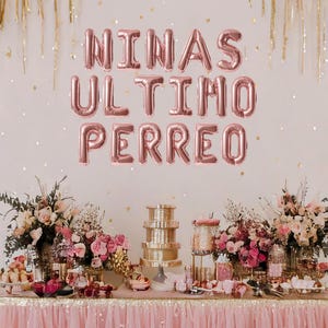 May include: A pink and gold party table setting with a banner that reads "NINAS ULTIMO PERREO". The table is decorated with flowers, candles, and other festive decorations.