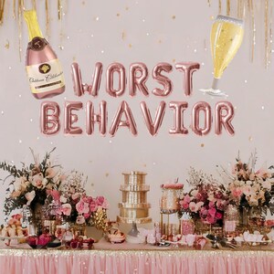May include: Rose gold foil balloons spelling "WORST BEHAVIOR" with a champagne bottle and glass balloon decorations. A table is set with a pink tablecloth and a dessert display with pink flowers.