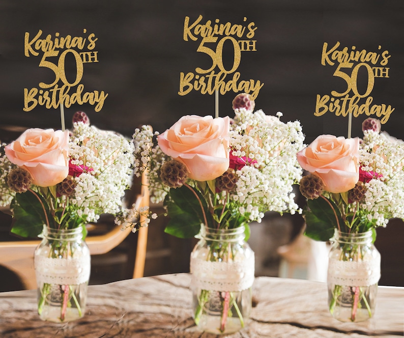 May include: Three gold glitter table centerpieces with the text "Karina's 50th Birthday" on each. The centerpieces are placed in mason jars with flowers.
