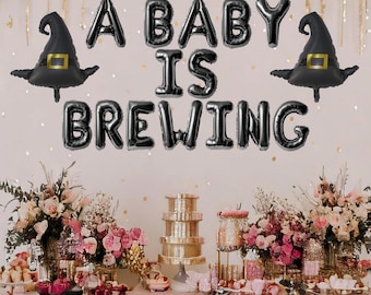 May include: A baby shower party decoration with black balloon letters spelling "A Baby Is Brewing" and two black witch hats. The letters and hats are on a pink and gold background with a table set up in front of the balloons.