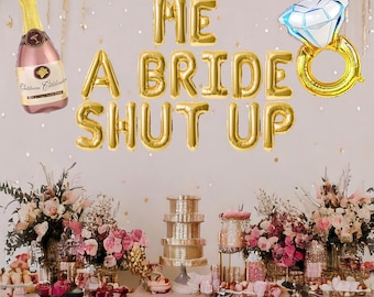 May include: Gold foil balloons spelling "HE A BRIDE SHUT UP" with a gold ring balloon with a blue diamond and a bottle of champagne. A table with a pink tablecloth and floral arrangements.
