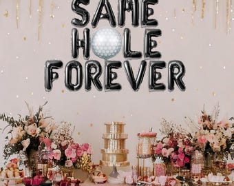 May include: Black balloon letters spelling "SAME HOLE FOREVER" with a white golf ball balloon in the center. The balloons are displayed on a pink and gold backdrop with a table set with flowers and desserts.