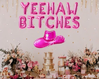 May include: Pink balloon letters spelling "Yeehaw Bitches" with a pink cowboy hat on a white background. The letters are hanging from the ceiling and there is a table set up in front of them with pink decorations.