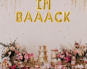 May include: A pink and gold dessert table with the words "I'm Baaack" spelled out in gold balloons. The table is decorated with pink flowers and gold accents.