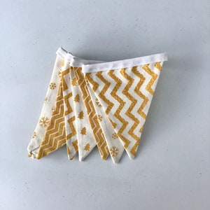 May include: A decorative banner composed of triangular flags. The flags display a white base with gold chevron, snowflake, and tree designs. A white band joins the flags, forming a garland. Suitable for seasonal or festive decorations.