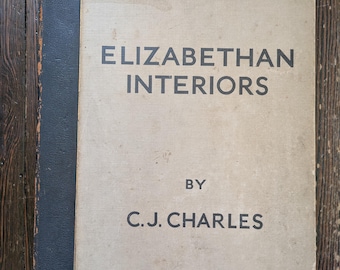 May include: A vintage book titled "ELIZABETHAN INTERIORS" by C.J. CHARLES. The book has a light tan cover with black text and a dark blue spine and corner accents. The book is on a wooden surface.