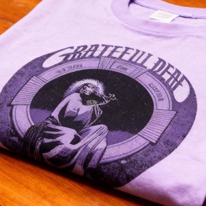 May include: A folded lilac t-shirt featuring a circular graphic with the text "Grateful Dead" and an illustration of a seated figure. The graphic is in shades of purple and black.