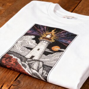 May include: White t-shirt featuring a detailed illustration of a lighthouse with a bright light, set against a sunset sky and ocean waves. The graphic is framed in a rectangular shape. The shirt is folded on a rustic wooden table.