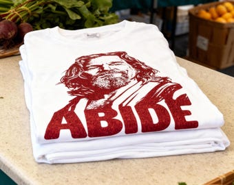 May include: White t-shirts stacked on a counter, each featuring a red graphic of a man's portrait and the word "ABIDE" in bold letters. The shirts are folded neatly, showcasing the design prominently.
