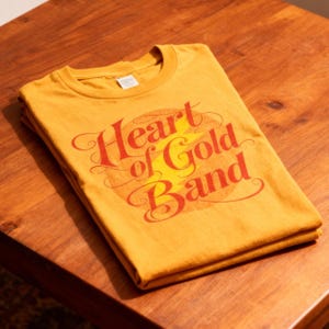 May include: Mustard yellow t-shirt with the text "Heart of Gold Band" in red script. The text is set over a yellow and orange sunburst design. The shirt is folded neatly on a wooden surface.