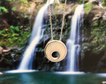 May include: A silver chain necklace with a white opal pendant in the shape of a circle with a hole in the center. The pendant is hanging in front of a blurry background of a waterfall.