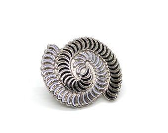 May include: A silver-toned spiral-shaped brooch with a detailed design. The brooch features alternating segments of black, white, and silver, creating a visually striking pattern. The pin is set against a white background.