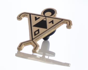 May include: Gold-colored metal pin with a black and white geometric design. The design features a stylized human figure with a circle for a head and a triangle for a body. The pin has a black rubber backing.