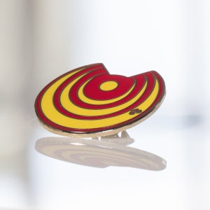 May include: A yellow and red enamel pin with a circular design. The pin has a gold finish and a safety clasp.
