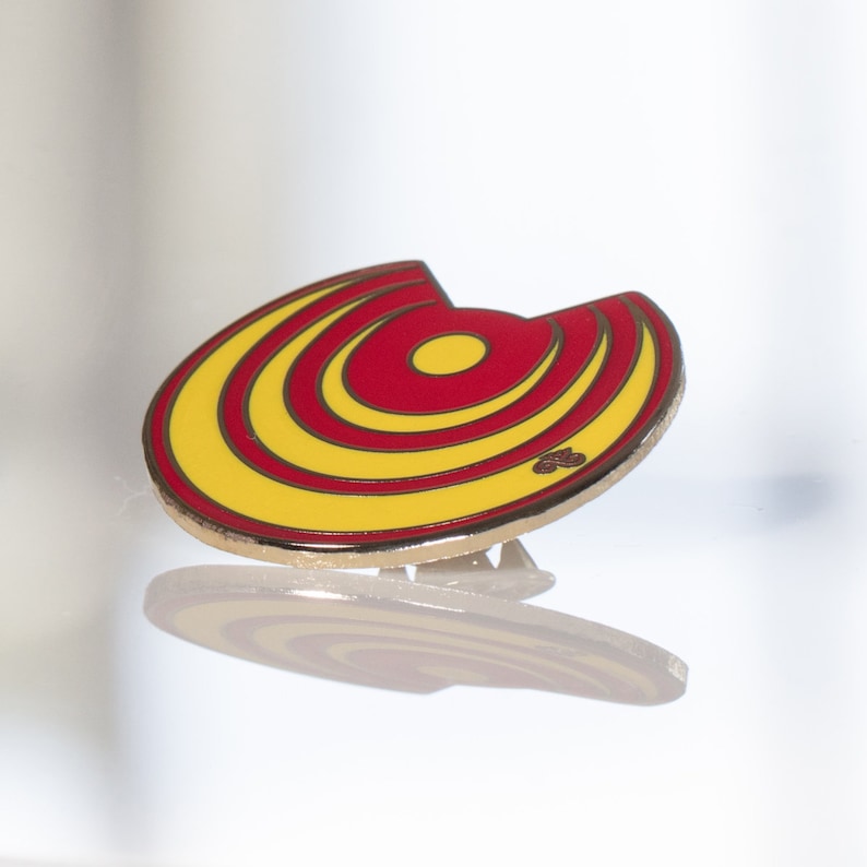 May include: A yellow and red enamel pin with a circular design. The pin has a gold finish and a safety clasp.