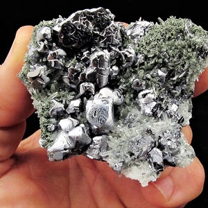 Könnte beinhalten: Ein Cluster aus silbrig-grauen metallischen Kristallen mit einer grünen Mineralmatrix. Die Kristalle sind scharf und kantig, mit einer reflektierenden Oberfläche.