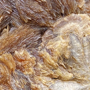 May include: Close-up of a brown and white mineral specimen with a rough, textured surface. The mineral has a layered, crystalline structure with a shimmery, iridescent sheen.