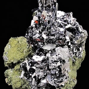 Könnte beinhalten: Nahaufnahme einer Mineralprobe mit einem Cluster aus metallischen, silberfarbenen Kristallen. Die Kristalle sind scharf definiert und reflektieren das Licht, vor dem Hintergrund grüner und weißer Mineralformationen. Der Hintergrund ist schwarz.