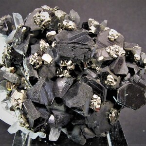 May include: A cluster of black, metallic, and shiny crystals with a few smaller, gold-colored crystals. The crystals are arranged in a random pattern and are set against a white background.