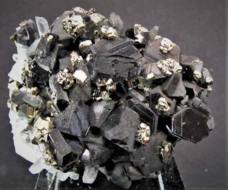 May include: A cluster of black, metallic, and shiny crystals with a few smaller, gold-colored crystals. The crystals are arranged in a random pattern and are set against a white background.