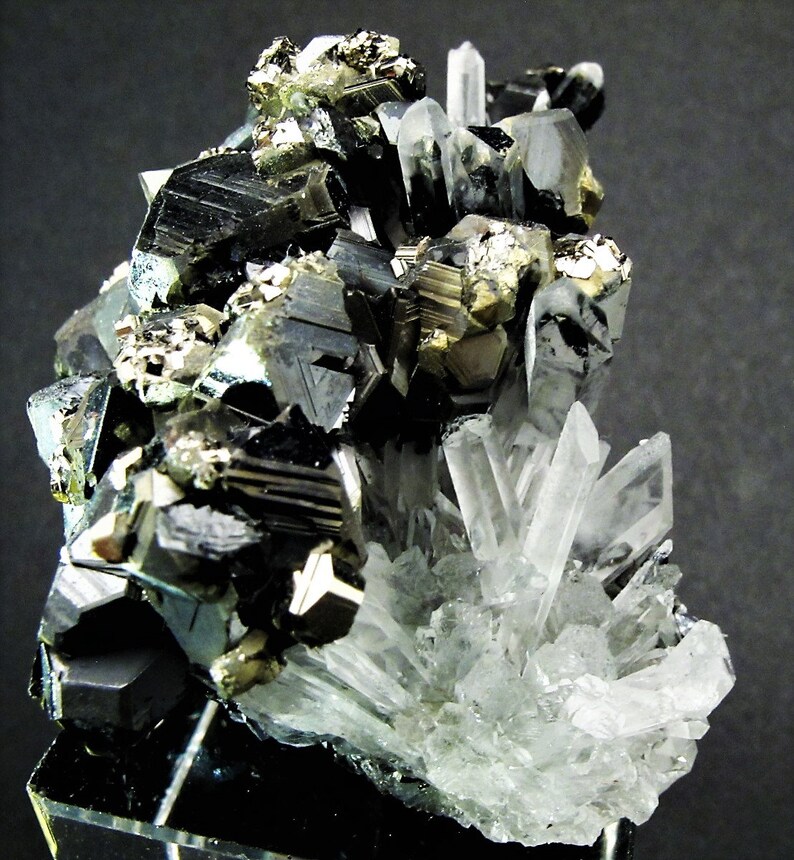 May include: A cluster of black, silver, and gold crystals on a black background. The crystals are arranged in a random pattern and are of varying sizes. The crystals are sharp and have a metallic sheen.