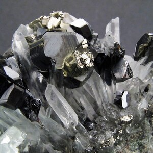May include: A cluster of black and silver crystals on a white background. The crystals are arranged in a random pattern and some are partially obscured by others. The crystals are sharp and have a metallic sheen.