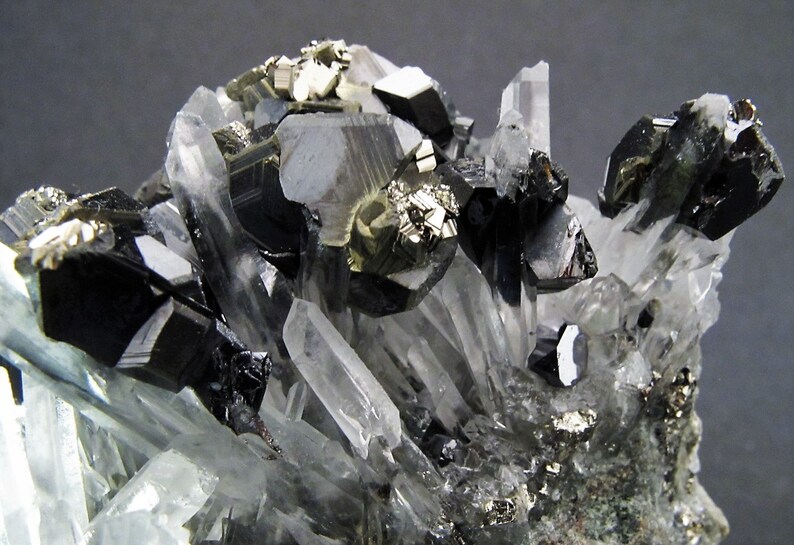 May include: A cluster of black and silver crystals on a white background. The crystals are arranged in a random pattern and some are partially obscured by others. The crystals are sharp and have a metallic sheen.