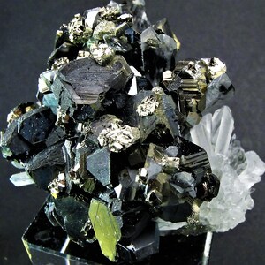 May include: A cluster of black and silver metallic crystals with a clear quartz crystal in the background. The crystals are arranged in a random pattern and have a shiny, reflective surface.