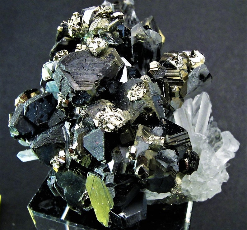 May include: A cluster of black and silver metallic crystals with a clear quartz crystal in the background. The crystals are arranged in a random pattern and have a shiny, reflective surface.