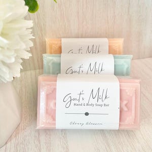 May include: Three bars of Goat's Milk Hand & Body Soap are stacked. The top bar is peach, the middle is mint green, and the bottom is pink. Each bar is wrapped in white paper with "Goat's Milk" printed on it. The bottom bar is labeled "Cherry Blossom."