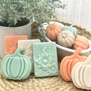 May include: A collection of decorative soaps in the shape of pumpkins and rectangular bars. The soaps are in various pastel colors, including orange, green, and cream. Some bars feature floral and leaf designs. The soaps are arranged on a woven mat.