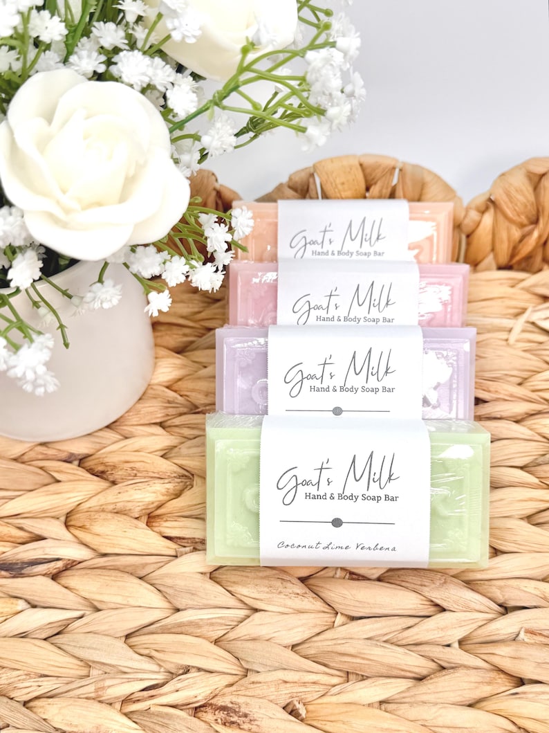 Puede incluir: Una pila de cuatro pastillas de jab&oacute;n rectangulares de leche de cabra para manos y cuerpo en colores naranja claro, rosa, morado y verde. Cada pastilla tiene una banda de papel blanco con el texto Goat's Milk Hand & Body Soap Bar. La pastilla verde inferior indica Coconut Lime Verbena.