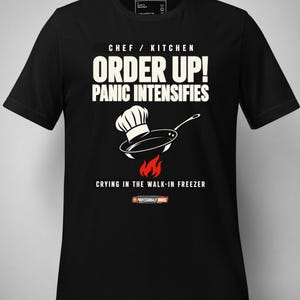 May include: A black short-sleeved t-shirt featuring a graphic with the text "CHEF / KITCHEN ORDER UP! PANIC INTENSIFIES" above an illustration of a chef hat, a frying pan, and red flames, with the text "CRYING IN THE WALK-IN FREEZER" below.