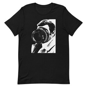 May include: Black and white graphic t-shirt featuring a man with a camera lens covering his face.