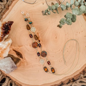 May include: A gold-toned eyeglass chain featuring a series of decorative beads in shades of orange, brown, and teal, interspersed with small gold-toned filigree circular charms and dragonfly-patterned round beads, ending in clear rubber loops.
