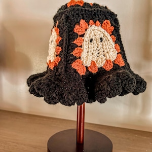 May include: A crocheted bucket hat featuring a black base with orange and cream-colored granny square motifs. Each square displays a central cream ghost figure with black eyes, surrounded by orange petals, finished with a ruffled black brim.