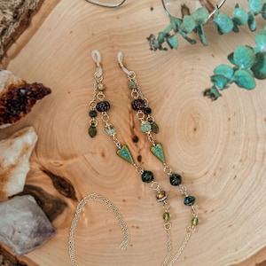 May include: A gold-toned metal eyeglass chain featuring a variety of decorative beads in shades of green, brown, and teal. The chain includes small leaf-shaped charms and round textured beads, connected by thin gold links with clear silicone loops.