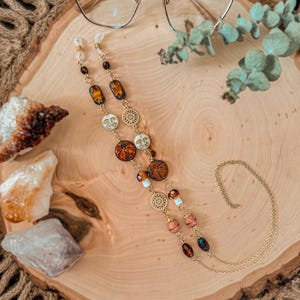 May include: A gold-toned eyeglass chain featuring a variety of decorative beads, including round charms with celestial face motifs, dragonfly patterns, and textured amber-colored glass beads, connected by delicate gold metal links and clear loops.