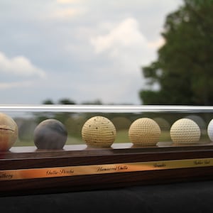 May include: A display case with six golf balls from different eras, including a feathery, gutta-percha, hammered gutta, bramble, rubber ball, and a modern ball. The case is made of wood with a brass plaque labeling each ball.